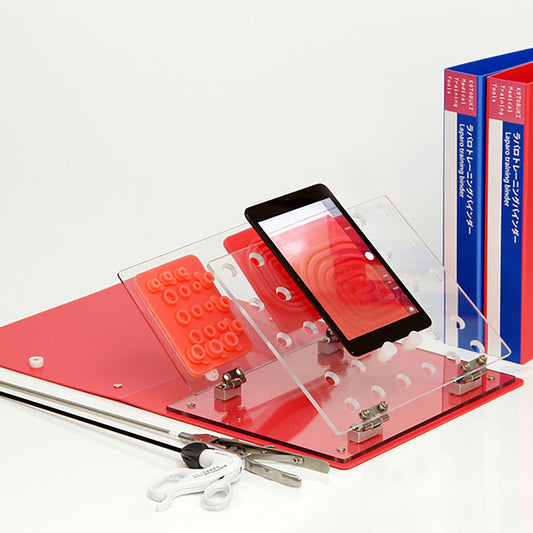 The Laparo Training Binder is laid flat on a desk. Both the clear laparoscopic wall and the setting base are tilted at a 45 degree angle to the left. An iPad rests on the laparoscopic wall in vertical position. Two silicon suture pads rest in vertical positions on the setting base. Two closed A4 binders stand upright to the side as for size comparisons.