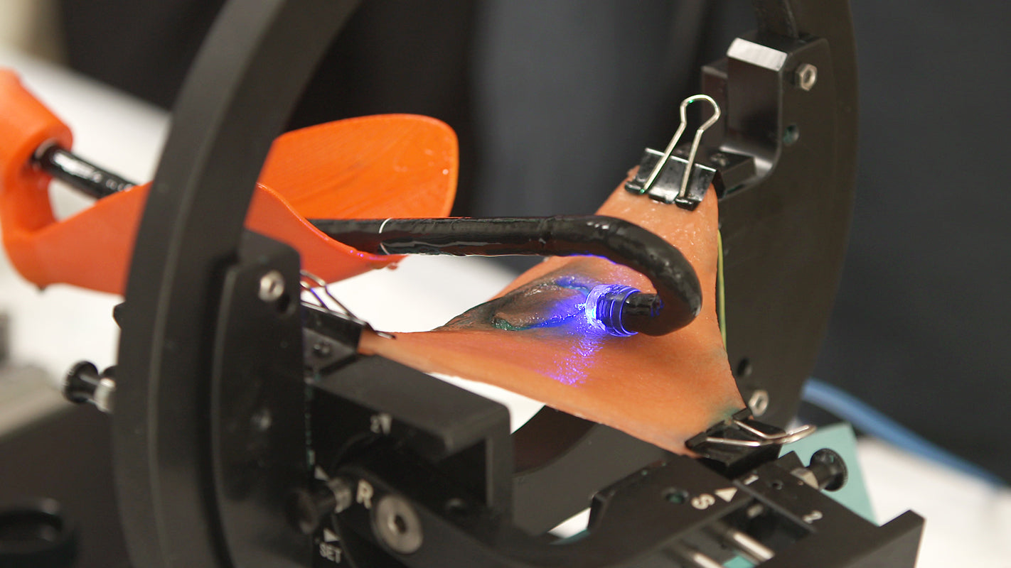 A blue-illuminated surgical endoscope sits above the synthetic lesion during an ESD training procedure. The training system is open-air, allowing a clear view of the endoscope and the electrosurgical incision. The gastric wall is simulated with an orange, plant-based tissue sheet injected with a blue lesion-raising solution.