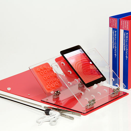 The Laparo Training Binder is laid flat on a desk. Both the clear laparoscopic wall and the setting base are tilted at a 45 degree angle to the left. An iPad rests on the laparoscopic wall in vertical position. Two silicon suture pads rest in vertical positions on the setting base. Two closed A4 binders stand upright to the side as for size comparisons.  