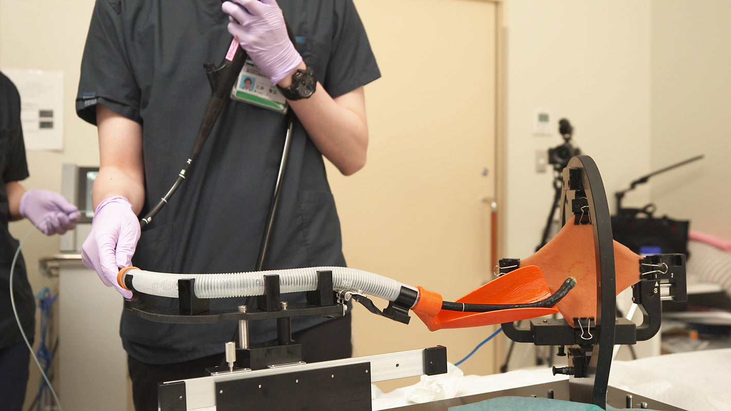 A GI fellow practices ESD using the G-Master training system. He maneuvers the endoscope through a clear tube and into the rotatable setting wheel, where an orange, plant-based tissue pad is stretched to represent the gastric wall.