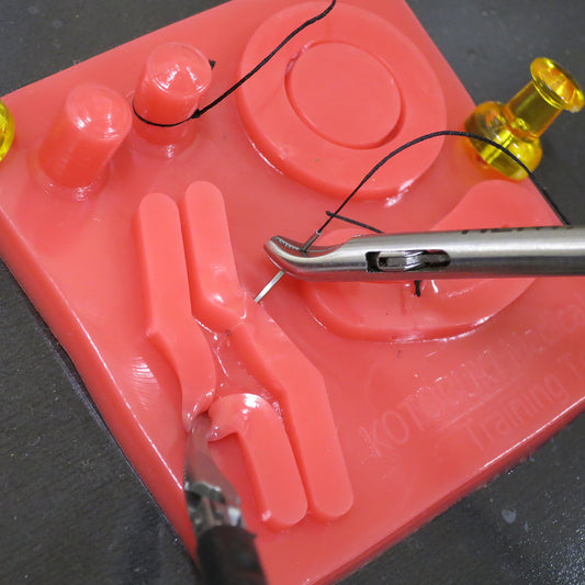 Laparoscopic ligature training performed on a miniature silicon suture pad. One trocar holds the thread while the other braces against the soft, red silicon. 
